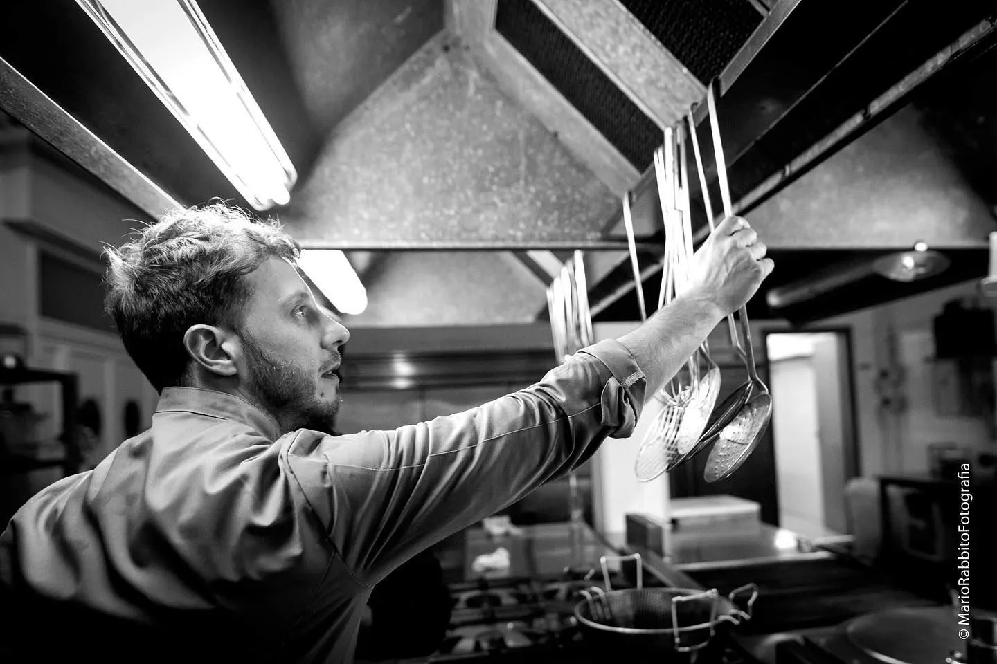 An editorial-style black and white image capturing a chef in a professional kitchen, reaching for cooking utensils. The image focuses on the action, highlighting the chef's hands and face, with kitchen equipment visible in the background.