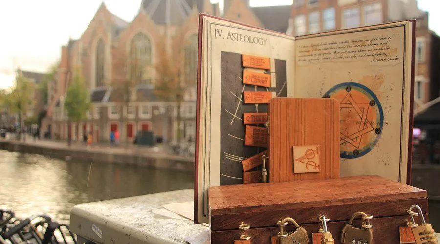 An open book displaying a chapter on 'Astrology' and a diagram, placed next to a small wooden box with ornate locks. The scene is set on a ledge or balcony, with a blurred background of a European cityscape featuring buildings and a canal.