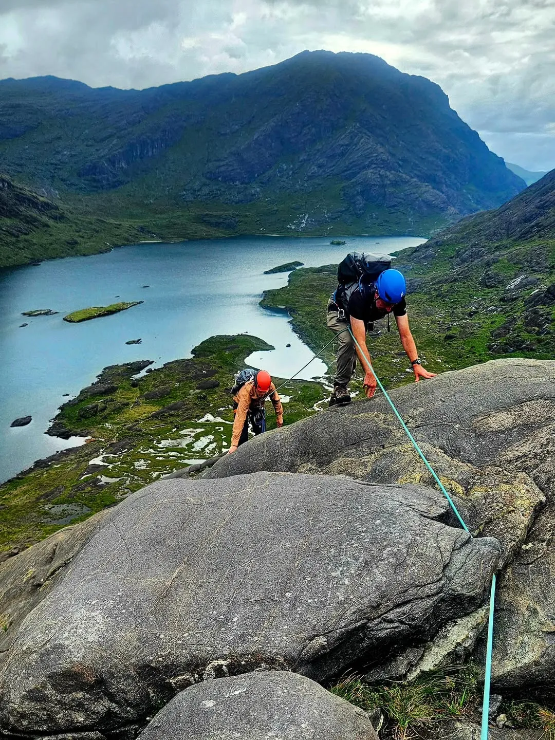Two people are seen rock climbing or scrambling up a rocky mountainside, with a beautiful blue lake and lush green valleys and distant mountains stretching behind them under a bright sky. The climbers are equipped with helmets, backpacks, and ropes, indicating a serious outdoor activity.