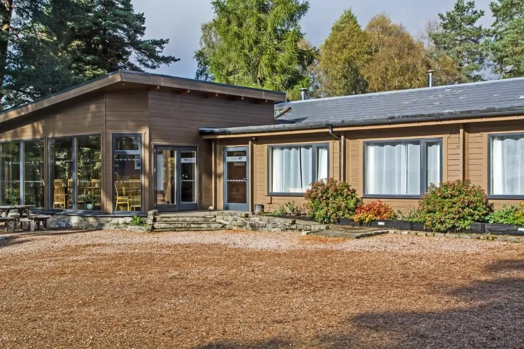 An exterior view of a modern brown wooden building with large windows and doors, surrounded by trees and a light brown gravel or mulch foreground.