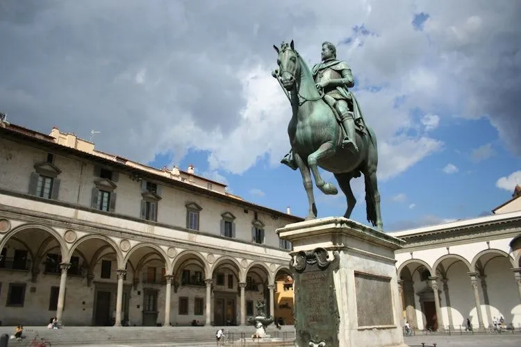 A well-composed editorial image depicting a prominent bronze equestrian statue in a sunlit public square, with an ornate historical building featuring arches in the background, set against a partly cloudy sky.