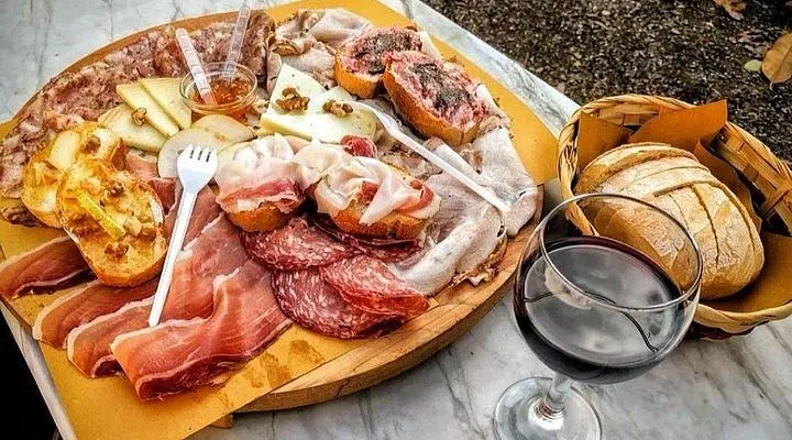 A well-composed, high-angle shot of a rustic charcuterie board laden with a variety of cured meats, cheeses, crostini, and condiments. A glass of red wine and a bread basket are visible in the background, set on a marble-textured surface.