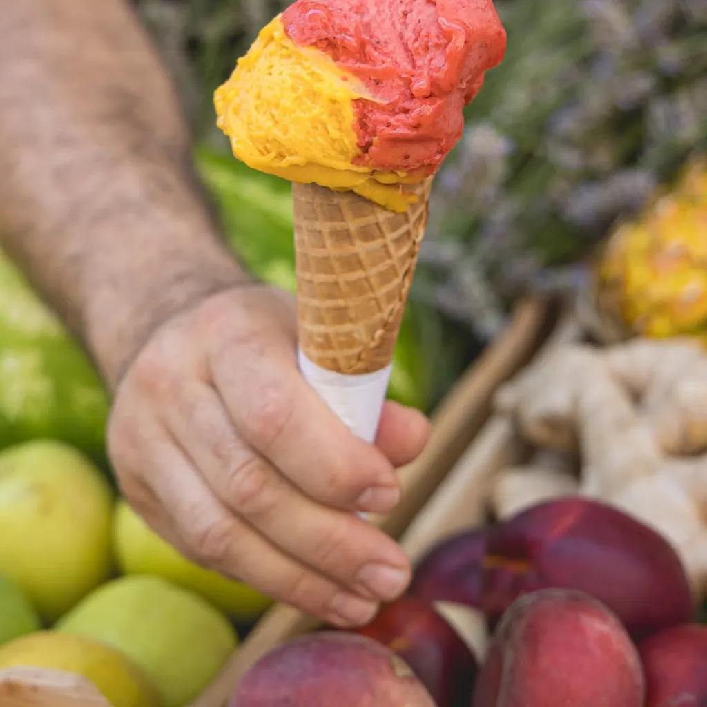 A vibrant image showcasing a two-scoop ice cream cone, held by a hand. The scoops feature distinct red/pink and yellow/orange colors, suggesting fruit flavors. The ice cream and cone are sharply in focus, while the background, which includes blurred elements like fruits and foliage, adds contextual depth without distracting.