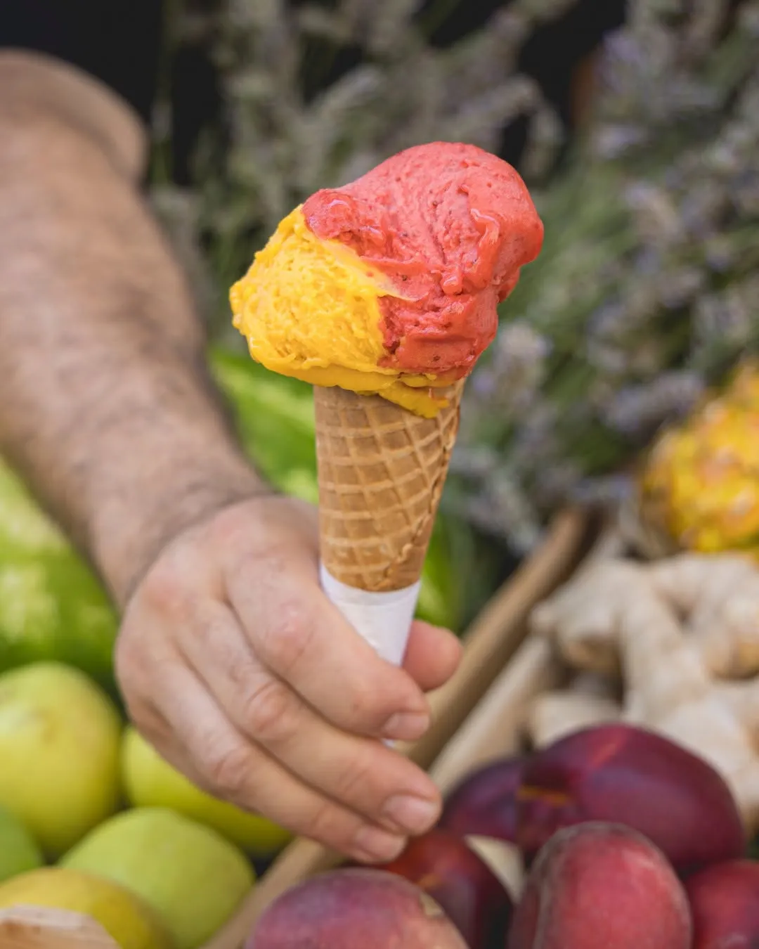 A vibrant image showcasing a two-scoop ice cream cone, held by a hand. The scoops feature distinct red/pink and yellow/orange colors, suggesting fruit flavors. The ice cream and cone are sharply in focus, while the background, which includes blurred elements like fruits and foliage, adds contextual depth without distracting.