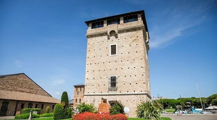 An imposing brick tower stands tall against a vibrant blue sky on a clear day, surrounded by well-maintained green spaces and colorful flowers. The image captures the architectural details of the tower, highlighting its historical significance.