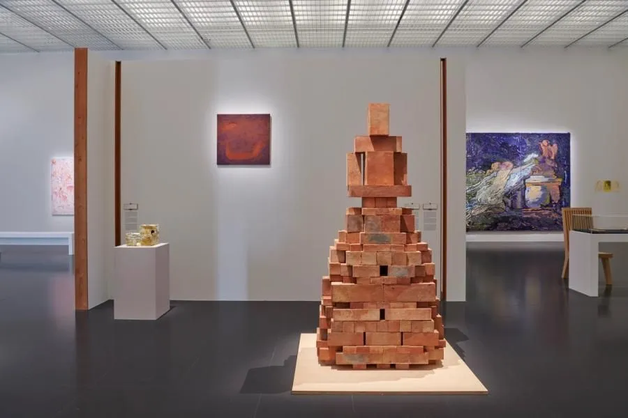 The image features a well-composed view of an art gallery, with a large brick sculpture serving as the central focal point. The lighting is even and effectively illuminates the various artworks, especially highlighting the texture of the brick sculpture. The image is sharp throughout, particularly on the main subject, and colors appear accurate and true to life for an indoor museum setting. The overall quality contributes to a highly photo-realistic representation of the scene.