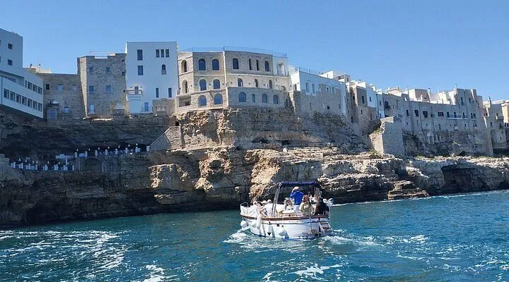 An editorial style image featuring a boat on the water in front of a coastal town built on cliffs under a clear blue sky. The scene depicts a sunny day with calm waters.