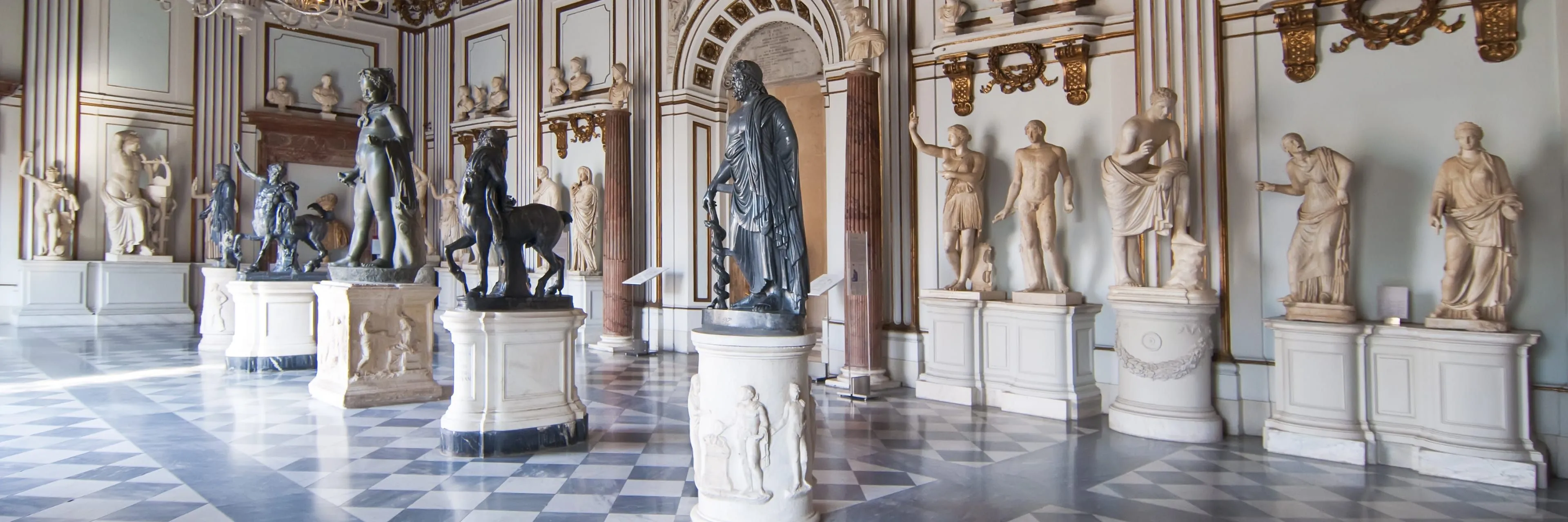 An interior shot of a grand museum hall featuring several classical sculptures, including a prominent bronze statue, set against a backdrop of ornate architectural details, columns, and a checkered floor.