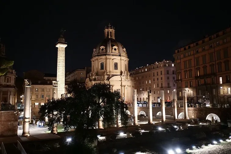 The image captures a beautiful night view of Roman monuments, including Trajan's Column and a prominent church dome. The composition effectively frames the illuminated buildings against the dark sky, creating a dramatic and appealing scene. The image is photo-realistic and the key architectural elements are in focus.