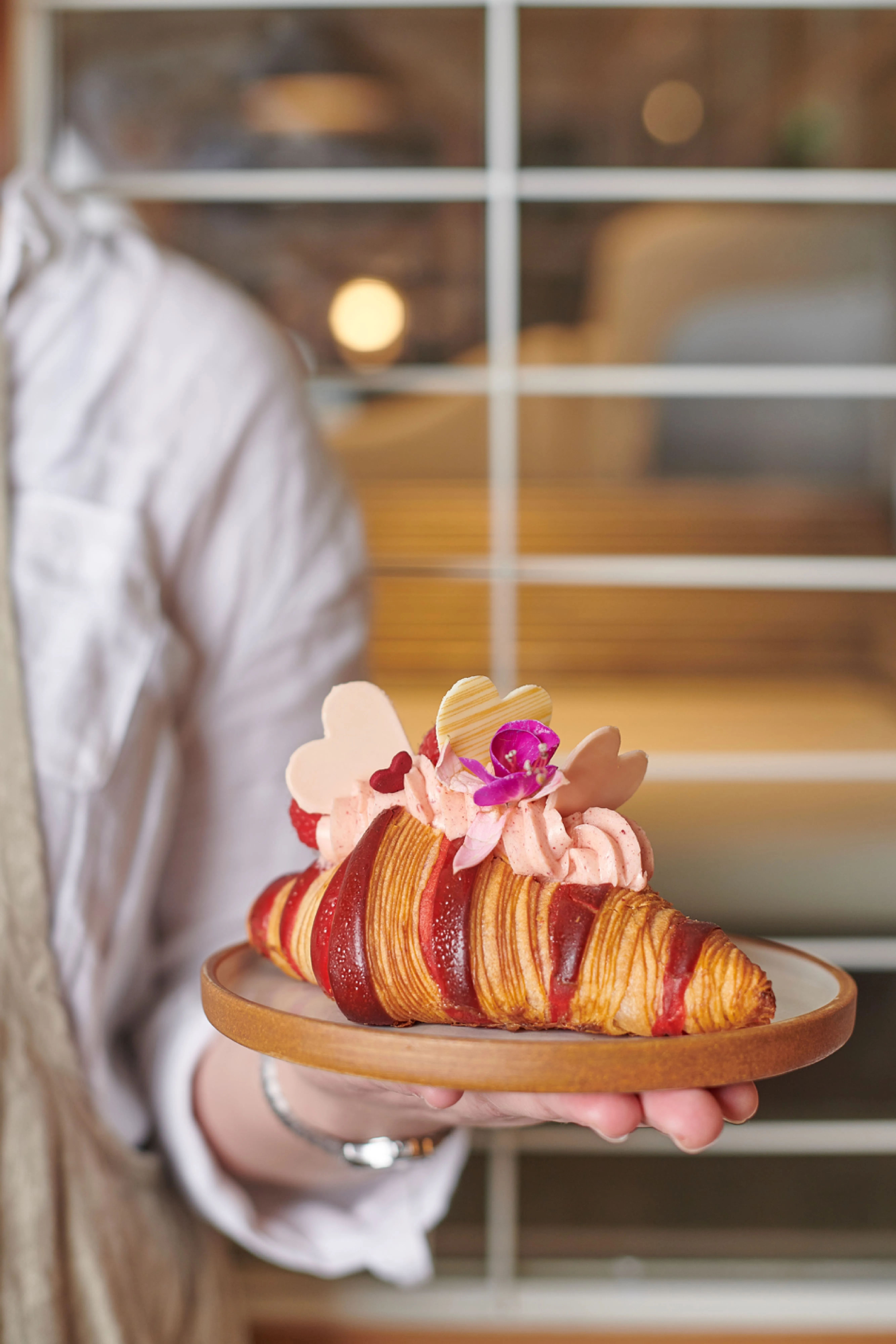 An exceptionally well-composed and visually appealing photograph of a decorated croissant. The pastry is the clear focal point, expertly framed, and held by a person in a way that enhances the presentation without distracting. The lighting is soft and even, highlighting the textures and vibrant colors of the red-striped croissant, pink cream, and heart-shaped decorations. The image is perfectly sharp, with all key elements in focus, contributing to its high photo-realism and overall professional quality.