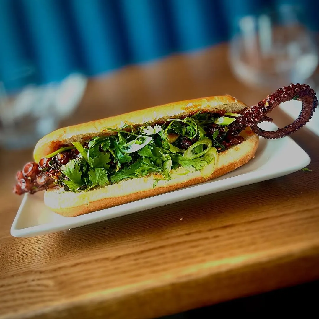 A close-up, professionally taken photograph of a gourmet sandwich, possibly a Banh Mi with octopus, presented on a white rectangular plate on a wooden surface. The sandwich is generously filled with fresh green herbs, and prominent octopus tentacles emerge from both ends. The background is softly blurred, enhancing the focus on the dish.