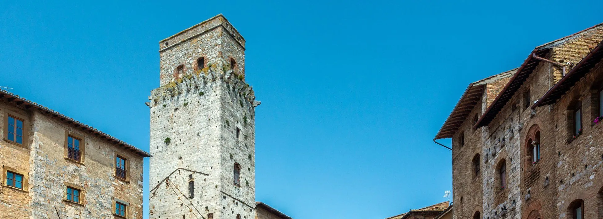 The image captures a tall stone tower against a clear blue sky. The tower, likely an old or medieval structure, fills the left side of the frame, showcasing its intricate stonework and windows. The lighting is even, highlighting the texture of the stone, and the blue sky provides a simple, uncluttered background.