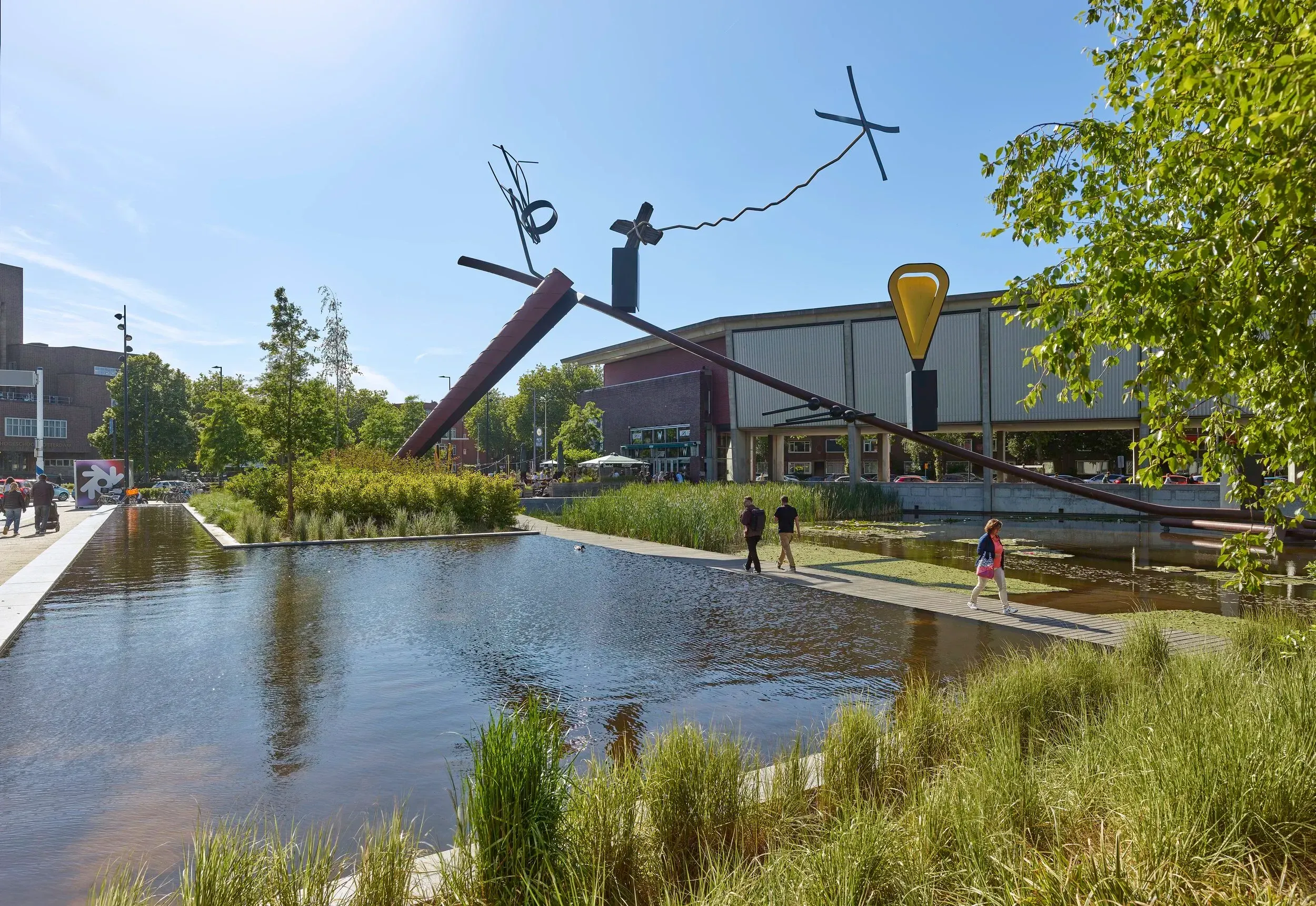 A well-lit, sharp, and color-accurate photograph of an urban park featuring a large abstract sculpture, a body of water, and a walkway under clear daylight. The image is photo-realistic and most elements are in focus.