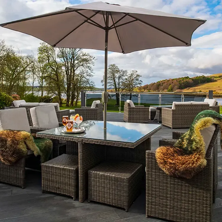 An outdoor patio furniture set with an umbrella and drinks on the table, overlooking a scenic lake and landscape under a cloudy sky.