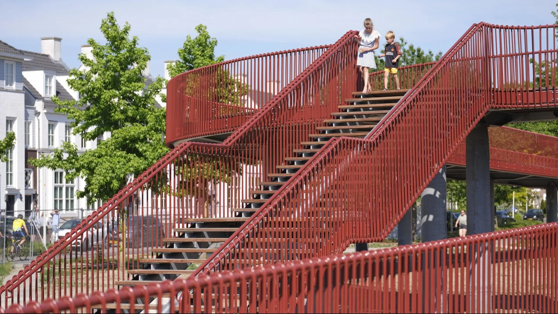The image features a visually appealing red architectural staircase or bridge, characterized by strong diagonal lines that effectively lead the eye. The composition is excellent, showcasing the structure against a clear sky and green foliage. Lighting is natural and even, contributing to vibrant and accurate colors. The photograph is sharp and highly photo-realistic, with the key architectural elements in clear focus. Two children are present on the stairs, providing a sense of scale without becoming the central focus.