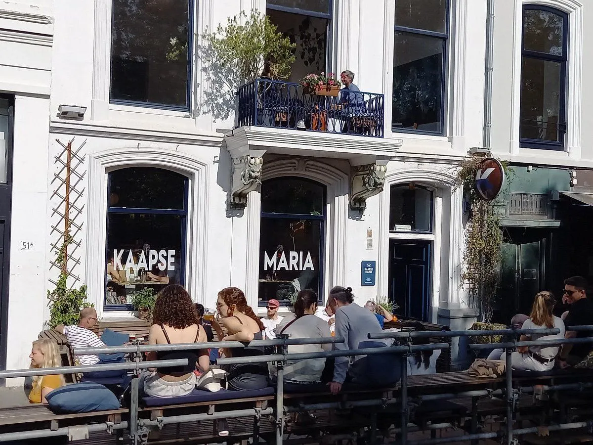 The image features strong natural lighting and accurate colors, lending to a highly photo-realistic appearance. Key elements like the building facade, window text, and people are generally in focus and sharp. However, the composition could be more refined, with some edge elements cut off, and the outdoor seating structure appears somewhat industrial, which might subtly detract from the overall aesthetic. Despite the strong sunlight, the image maintains good exposure, although some areas exhibit strong highlights and shadows.