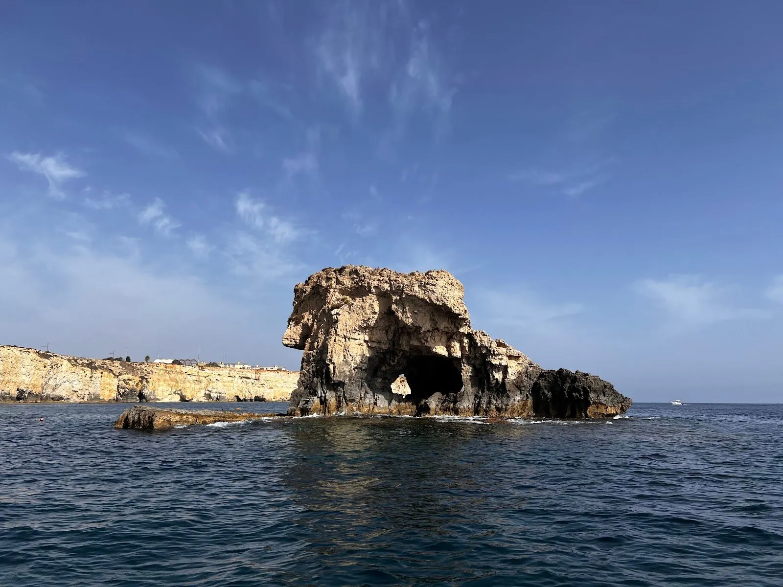 A clear, well-lit, and realistic photograph of a large rock formation with a natural arch, situated in blue ocean water under a clear sky. In the background, a distant coastline is visible.