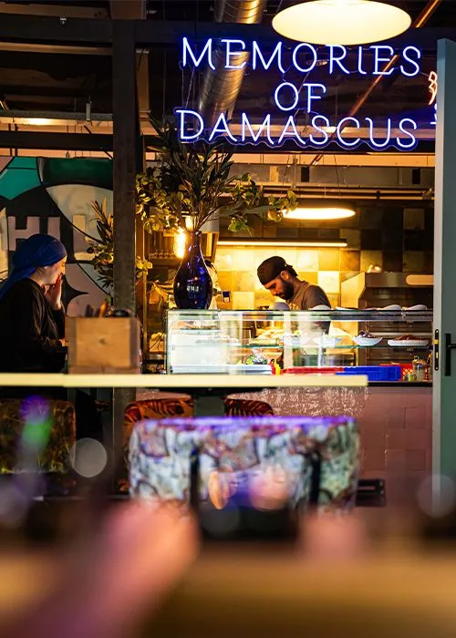 An editorial image capturing a candid moment inside a vibrant restaurant or bar. It features a woman seated at a counter in the foreground, with a chef actively working in the kitchen area behind her. The scene is characterized by atmospheric artificial lighting, including a prominent blue neon sign spelling 'OF DAMASCUS' and warm kitchen lights, creating a dynamic and inviting ambiance.