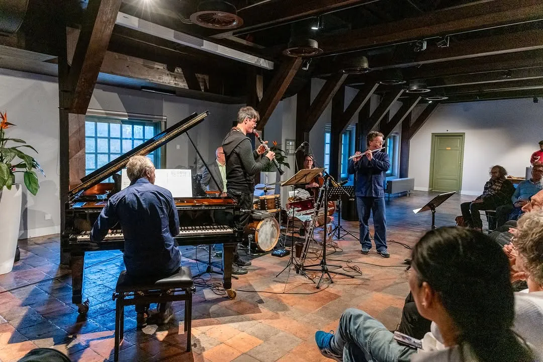 The image successfully captures a live musical performance indoors, featuring a group of musicians playing various instruments including a piano, clarinet, and flute. The scene is realistic, sharp, and the colors appear accurate. The composition effectively places the musicians as the central focus, providing a sense of being part of the audience. The lighting is generally good, though there are areas of strong contrast from the windows and some uneven shadows.