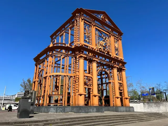 The image features a well-composed and sharply focused photograph of an intricate, orange architectural sculpture set against a vibrant blue sky. Excellent natural lighting highlights the structure's details and ensures color accuracy, contributing to its high photo-realistic quality.