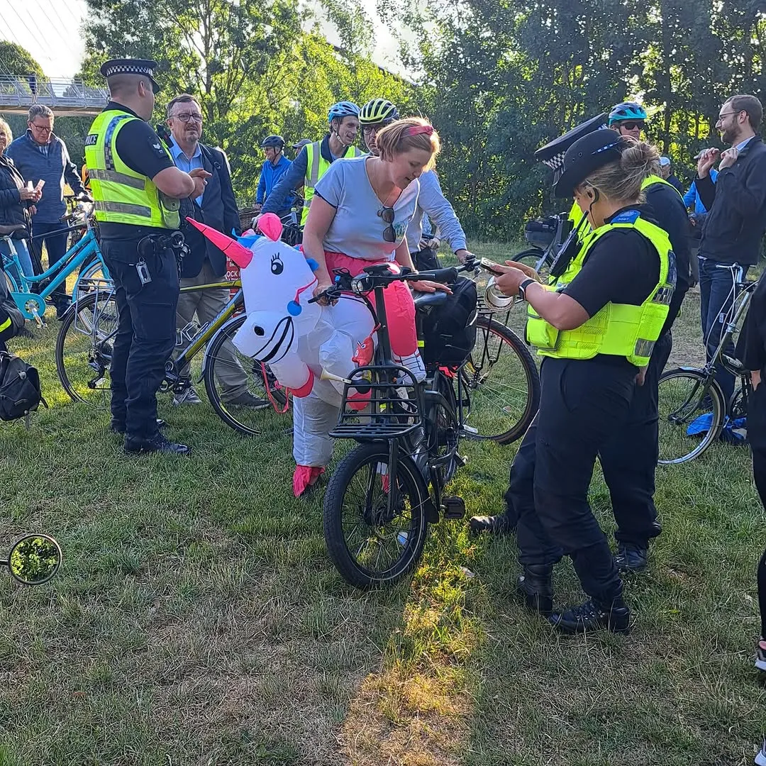 A group of people, including police officers in high-visibility vests, are gathered outdoors with bicycles on a grassy area. One individual is notably wearing an inflatable unicorn costume while on a bike, suggesting a lighthearted community event.