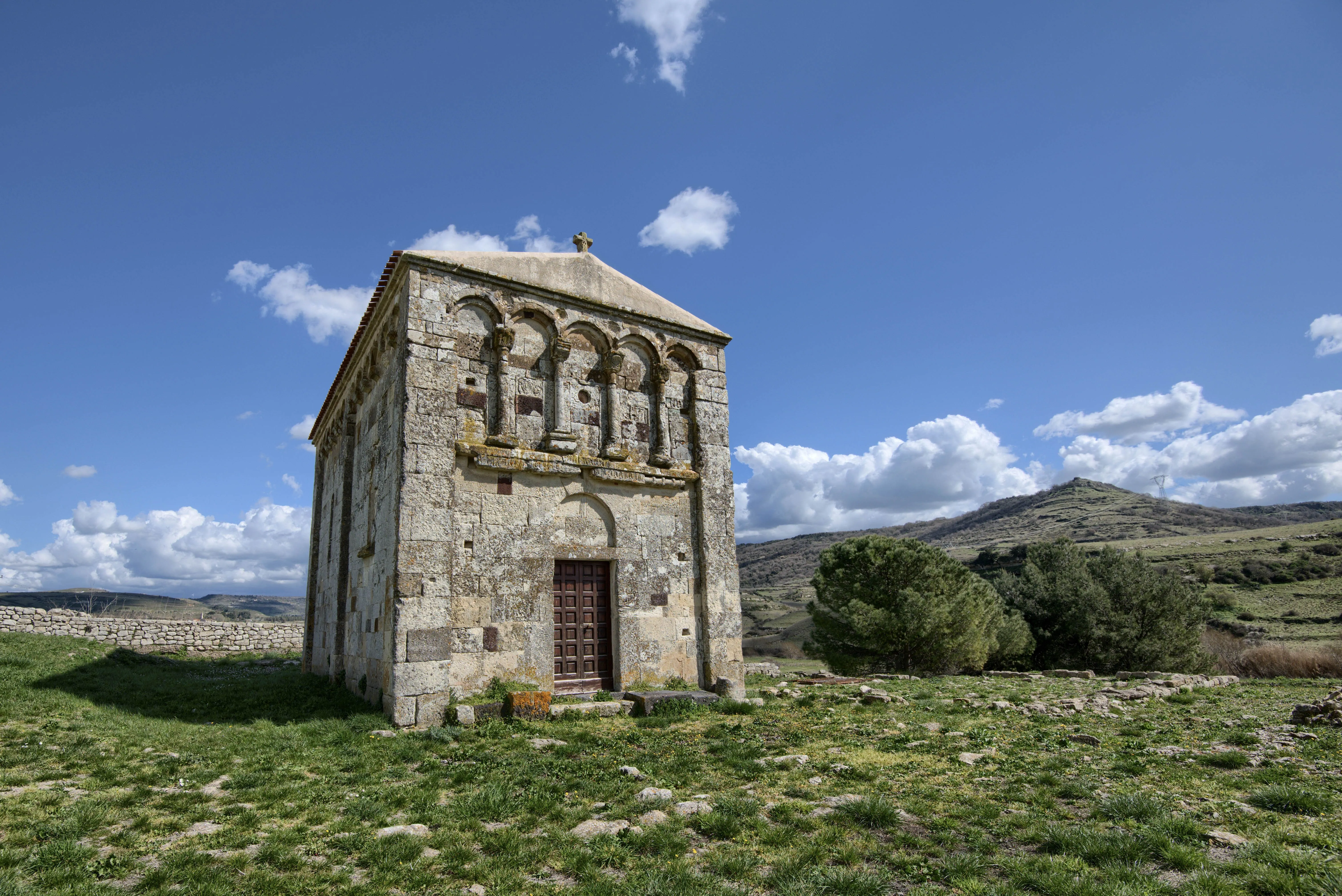 The image is well-lit with natural daylight, showcasing a stone building under a clear blue sky with white clouds. The main subject, the building, is sharp and in clear focus, with good detail visible in its stone texture. Colors appear natural and accurate, contributing to a strong photo-realistic quality. The composition is generally good, effectively presenting the building within its landscape.