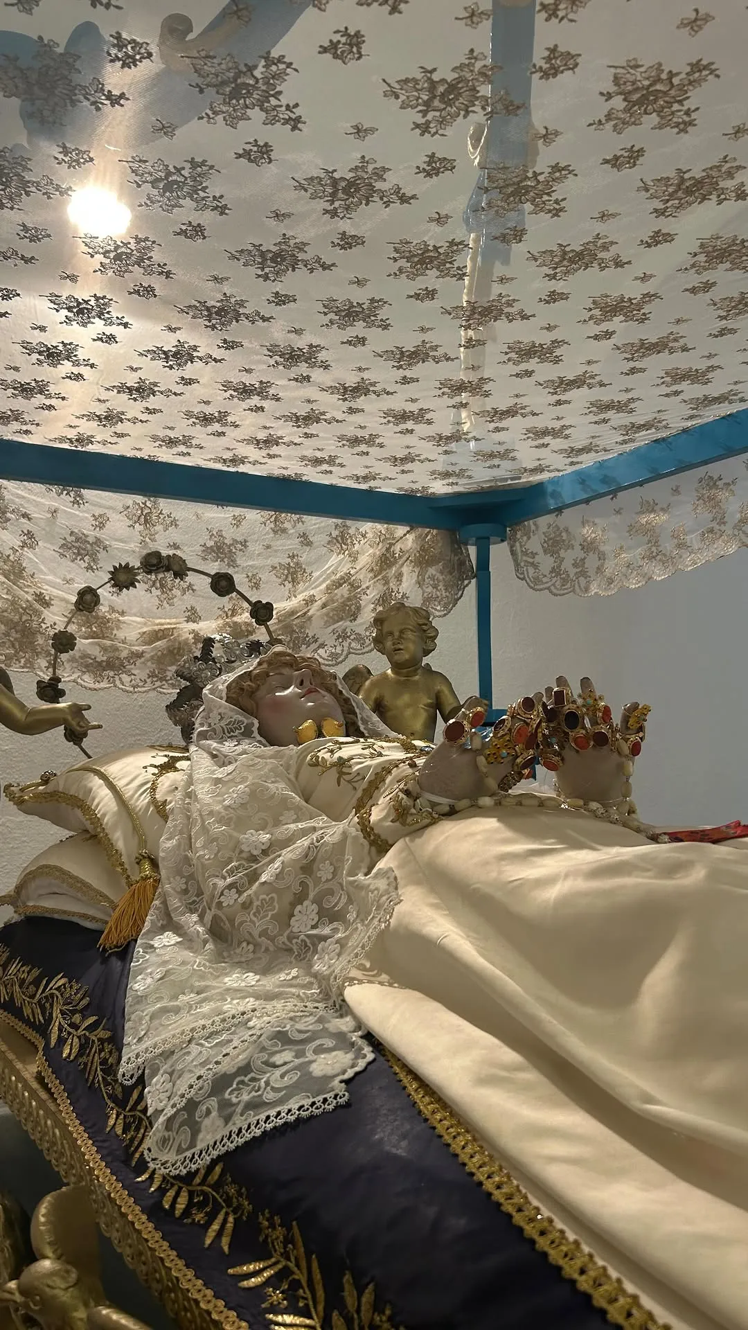 An elaborately dressed historical or religious effigy, possibly a saint, lying in repose under a patterned canopy, adorned with intricate lace, golden details, and a jeweled crown.