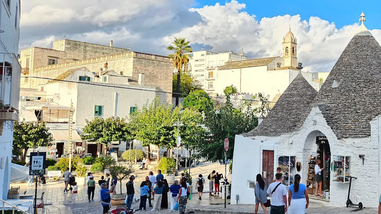 An editorial style image depicting a lively town square or street scene under a partly cloudy sky. The scene features traditional white buildings, green trees, and numerous people going about their day. A distinct trullo roof is visible in the foreground on the right, suggesting a location like Alberobello in Italy.