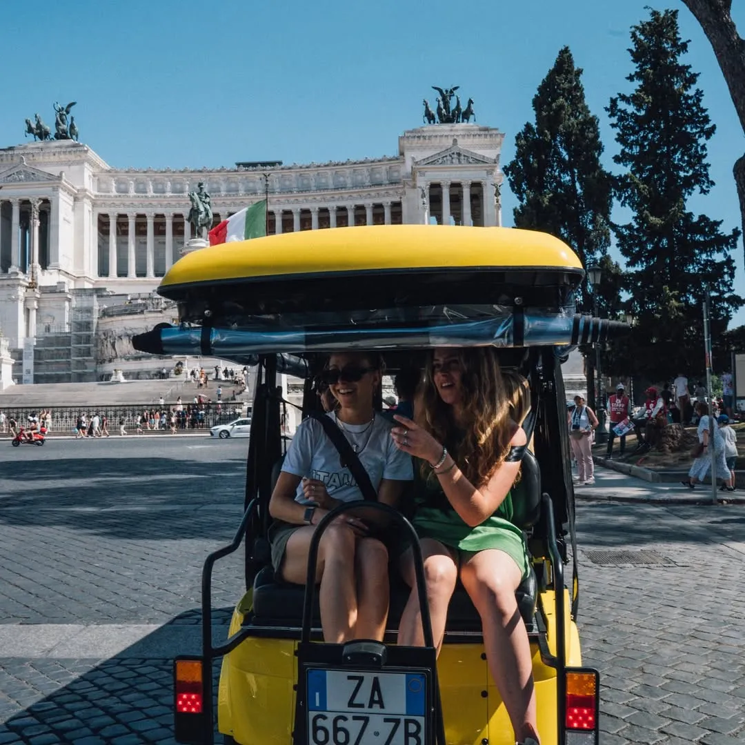 Two people are seen riding in a yellow open-air vehicle, with a large, ornate white stone monument visible in the background under a clear blue sky. The scene appears to be a sunny day in a city, possibly Rome, capturing a moment of sightseeing.