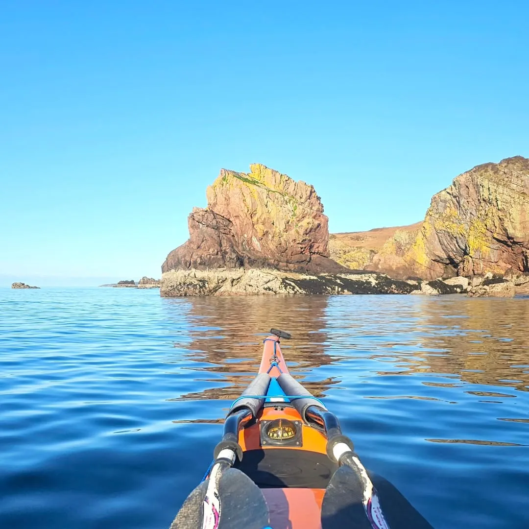 The image boasts excellent quality, characterized by vibrant and accurate colors, natural lighting, and overall sharpness. The composition effectively places the viewer in the kayak, creating an immersive first-person perspective of the vast blue waters and impressive rock formations. Both the foreground elements (kayak) and the distant landscape are in sharp focus, contributing to the photo-realistic and clear presentation of the scene. The image is well-lit, highlighting the details of the rocks and the clarity of the water without harsh shadows or overexposure.