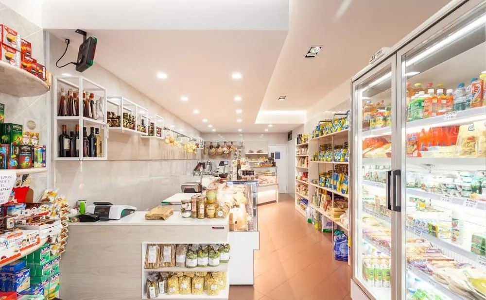 An interior view of a well-stocked and brightly lit deli or grocery store, featuring a counter with display cases and shelves filled with various food products.