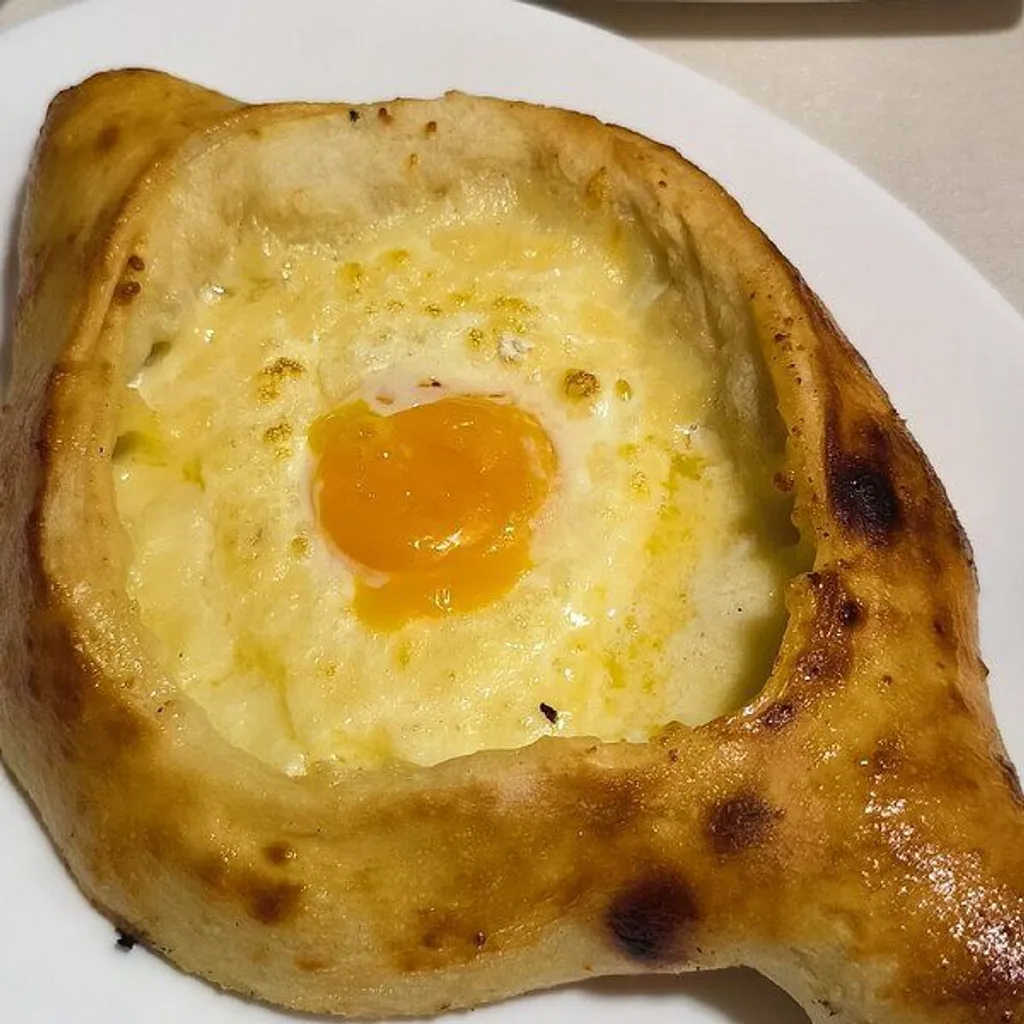 A close-up shot of a traditional Georgian Khachapuri Adjaruli, featuring a golden-brown baked dough boat filled with melted cheese and topped with a bright orange egg yolk.