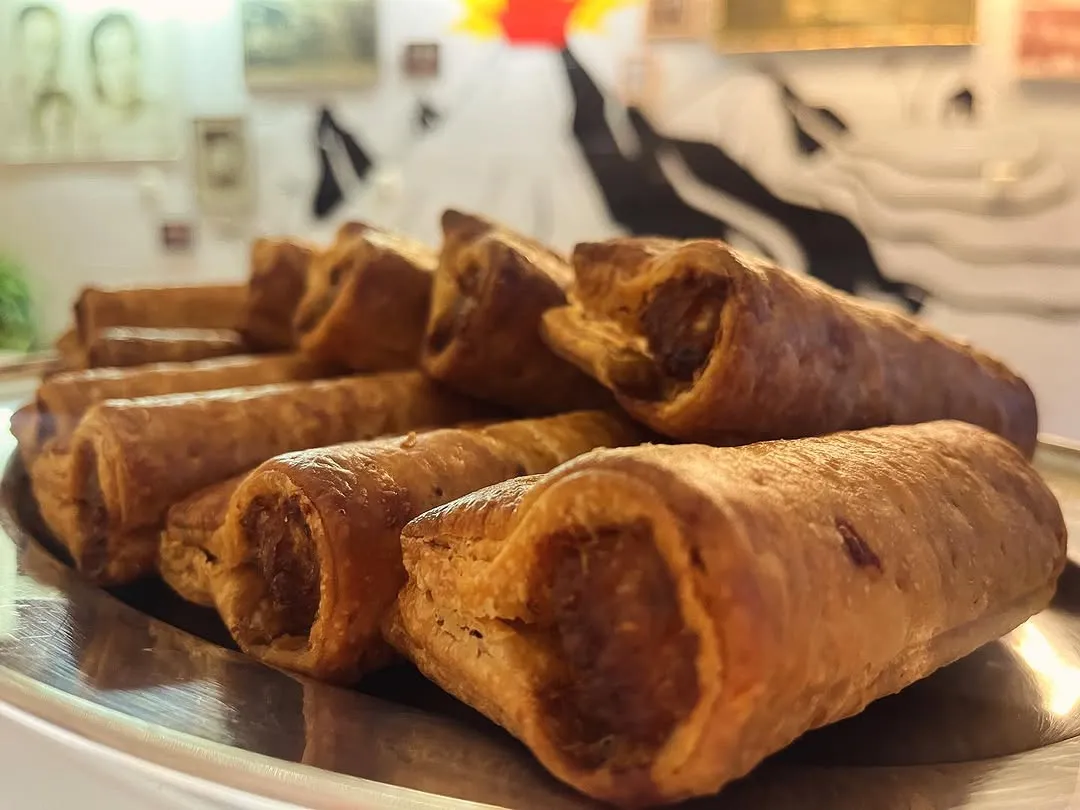 A close-up, eye-level shot of several golden-brown sausage rolls piled on a tray. The foreground items are sharply in focus, showcasing the flaky pastry texture. The background is blurred, featuring abstract patterns and faint hints of artwork.