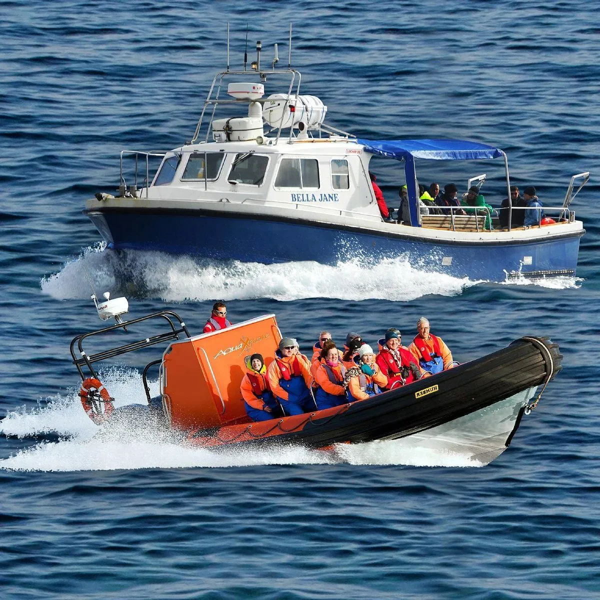 A dynamic scene featuring two boats, a larger blue and white cabin cruiser and a smaller orange and black rigid inflatable boat (RIB), moving across blue water, creating wakes. Both boats are occupied by passengers.