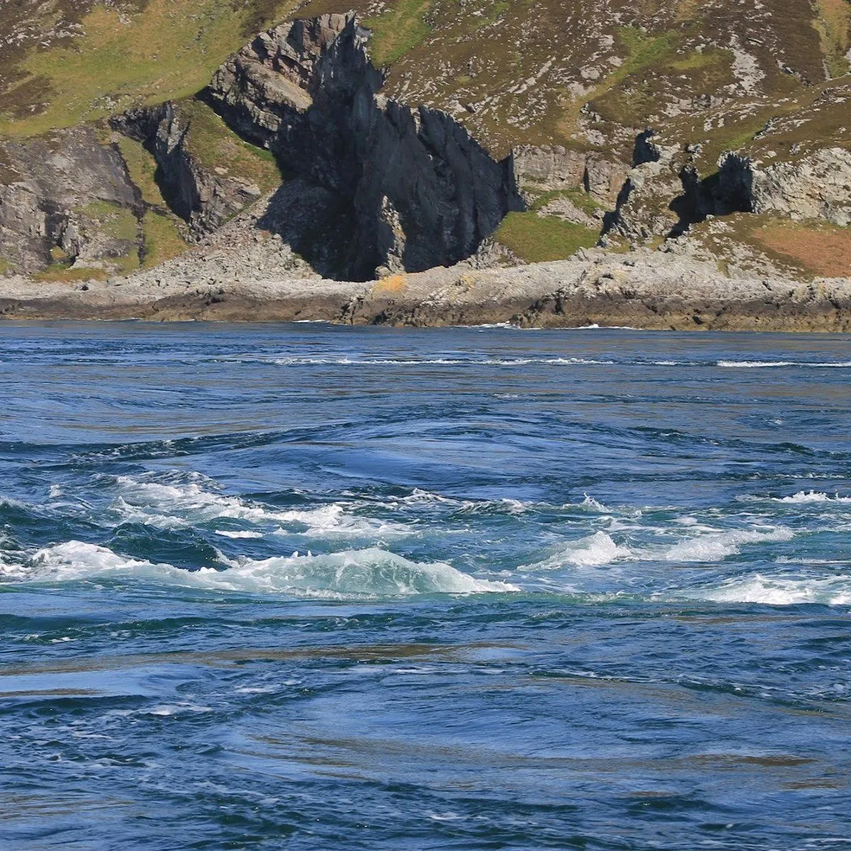 A dynamic view of turbulent blue water with white foam in the foreground, set against a backdrop of rugged, rocky cliffs covered with patches of green vegetation under bright daylight.