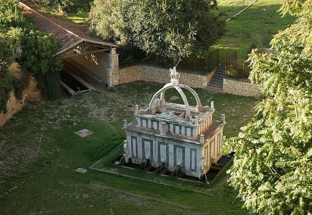 An aerial view of a historic stone fountain with multiple spouts and an ornate dome, situated in a green, grassy area. An old, covered washhouse structure is visible to the left, and trees and stone walls frame the scene.