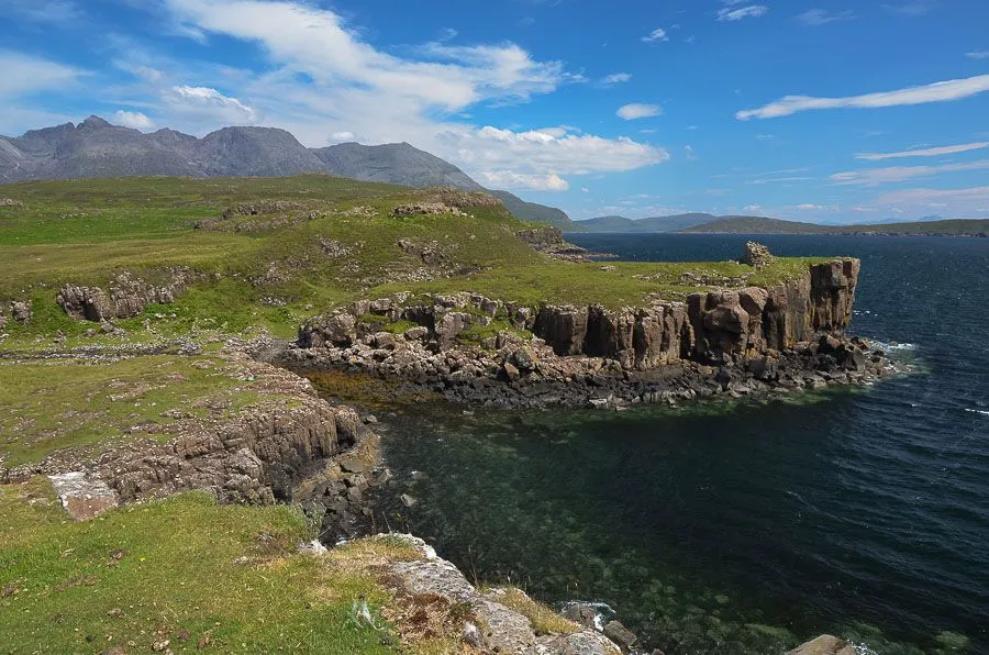 A breathtaking elevated view of rugged coastal cliffs meeting a calm, clear body of water, with rolling green hills extending towards distant mountains under a bright blue sky with wispy clouds.