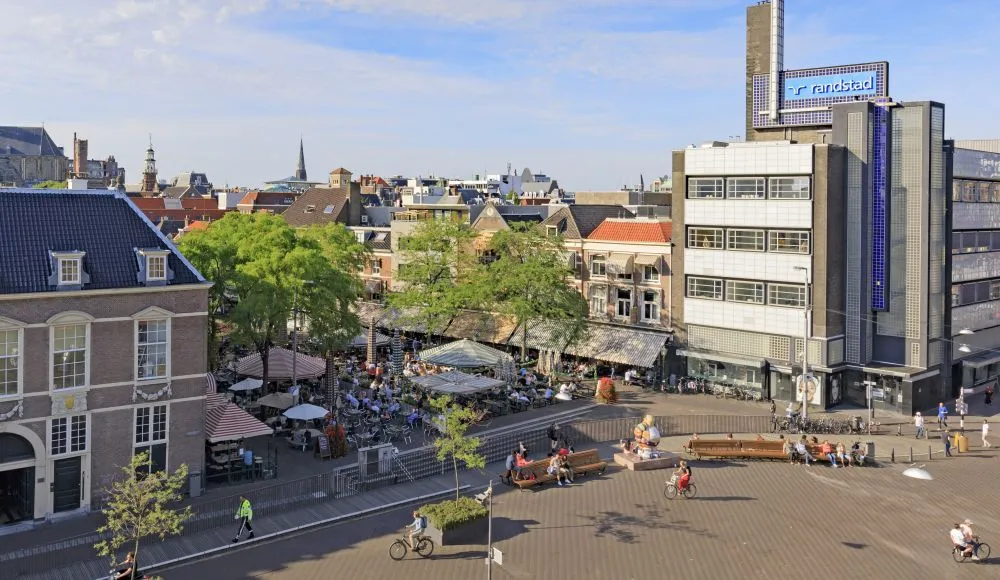 An aerial view of a lively city square featuring outdoor cafes, people walking and cycling, and historic buildings under a clear blue sky. The scene captures typical urban life during the day.