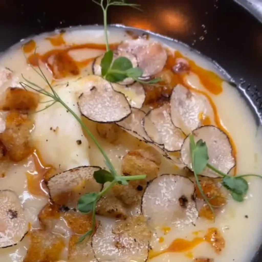 A close-up shot of a creamy, light-colored soup or puree, generously garnished with thin slices of truffle, golden-brown croutons, and fresh green microgreens. There are also hints of an orange-red oil or sauce drizzled on top, adding a touch of color.