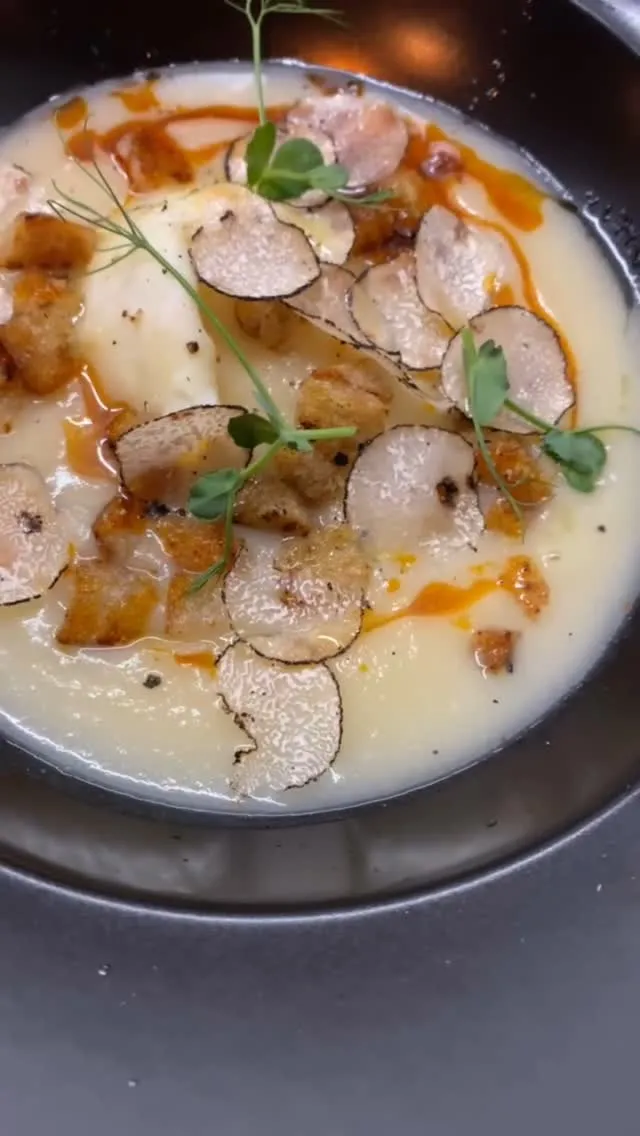 A close-up shot of a creamy, light-colored soup or puree, generously garnished with thin slices of truffle, golden-brown croutons, and fresh green microgreens. There are also hints of an orange-red oil or sauce drizzled on top, adding a touch of color.