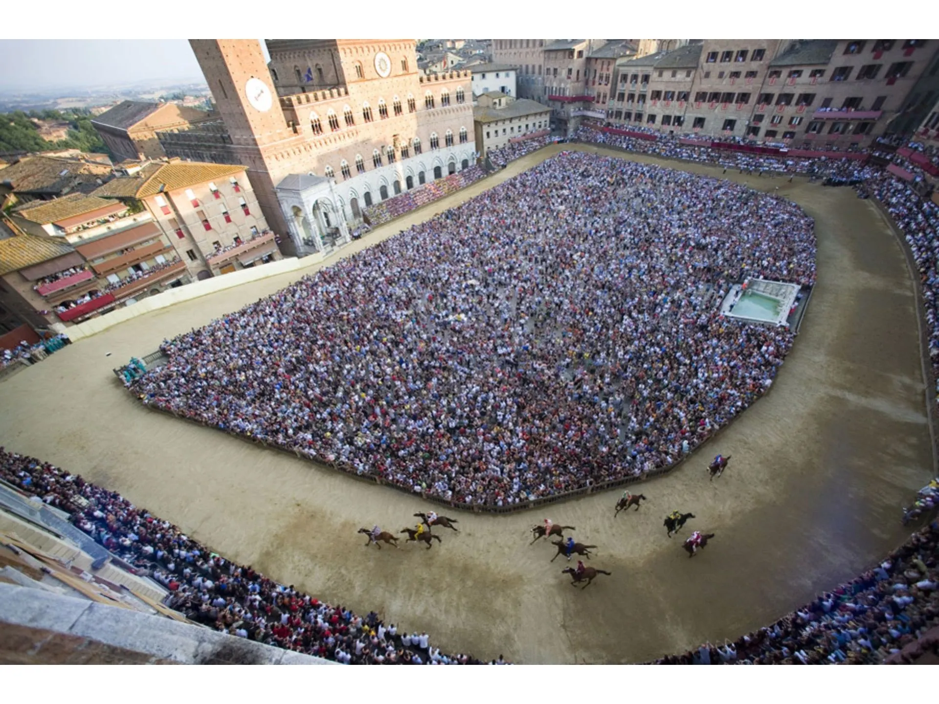 This image captures the excitement of the Palio di Siena from an aerial perspective, providing a comprehensive view of the horse race, the massive crowd, and the historic Piazza del Campo. The natural lighting is excellent, contributing to vibrant and accurate colors. Key elements like the horses and the track are sharp, and the overall photo-realism is high, effectively conveying the scale and atmosphere of the event.