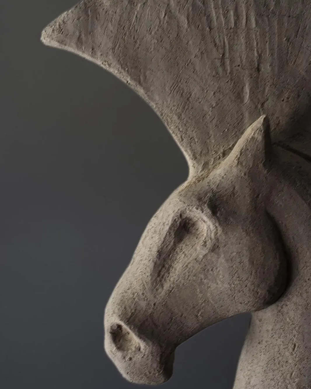 A close-up photograph of a sculpture, likely depicting a horse's head and neck, made from a textured, grey material resembling stone or clay. The lighting accentuates the sculpture's form and surface details against a plain, dark background.