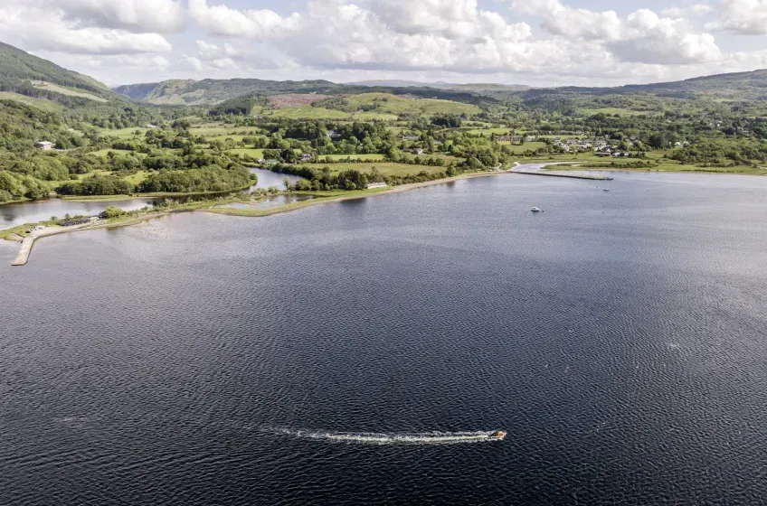 An aerial view captures a large expanse of deep blue water with a small boat creating a white wake, bordered by a green, hilly landscape dotted with trees and structures under a bright, cloudy sky.