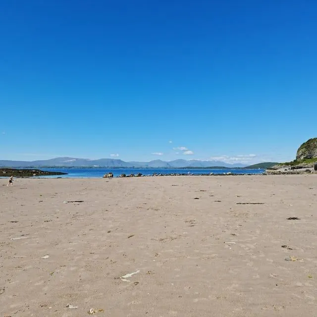 The image is well-lit with natural sunlight, showcasing accurate colors of the sky, sea, and sand. It is sharp and photo-realistic, with all key elements in focus. The composition is generally good, capturing a wide expanse of the beach and distant mountains.