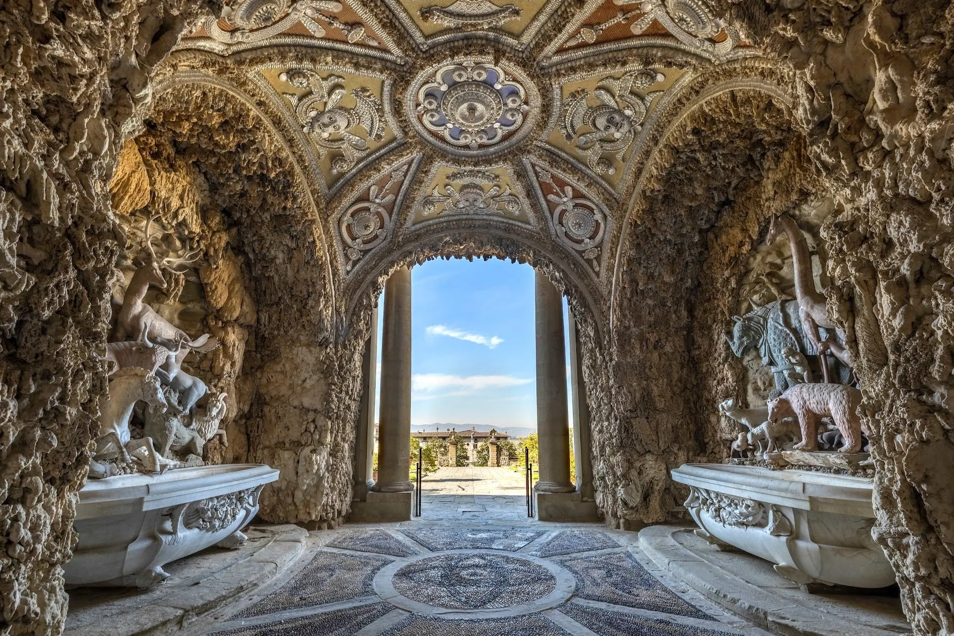 An interior perspective of an ornate architectural passage or grotto, featuring a highly detailed vaulted ceiling and rough-hewn stone walls with sculpted elements. The view looks out through a grand archway to a bright blue sky and a distant landscape with classical architecture. A patterned mosaic floor is visible in the foreground.