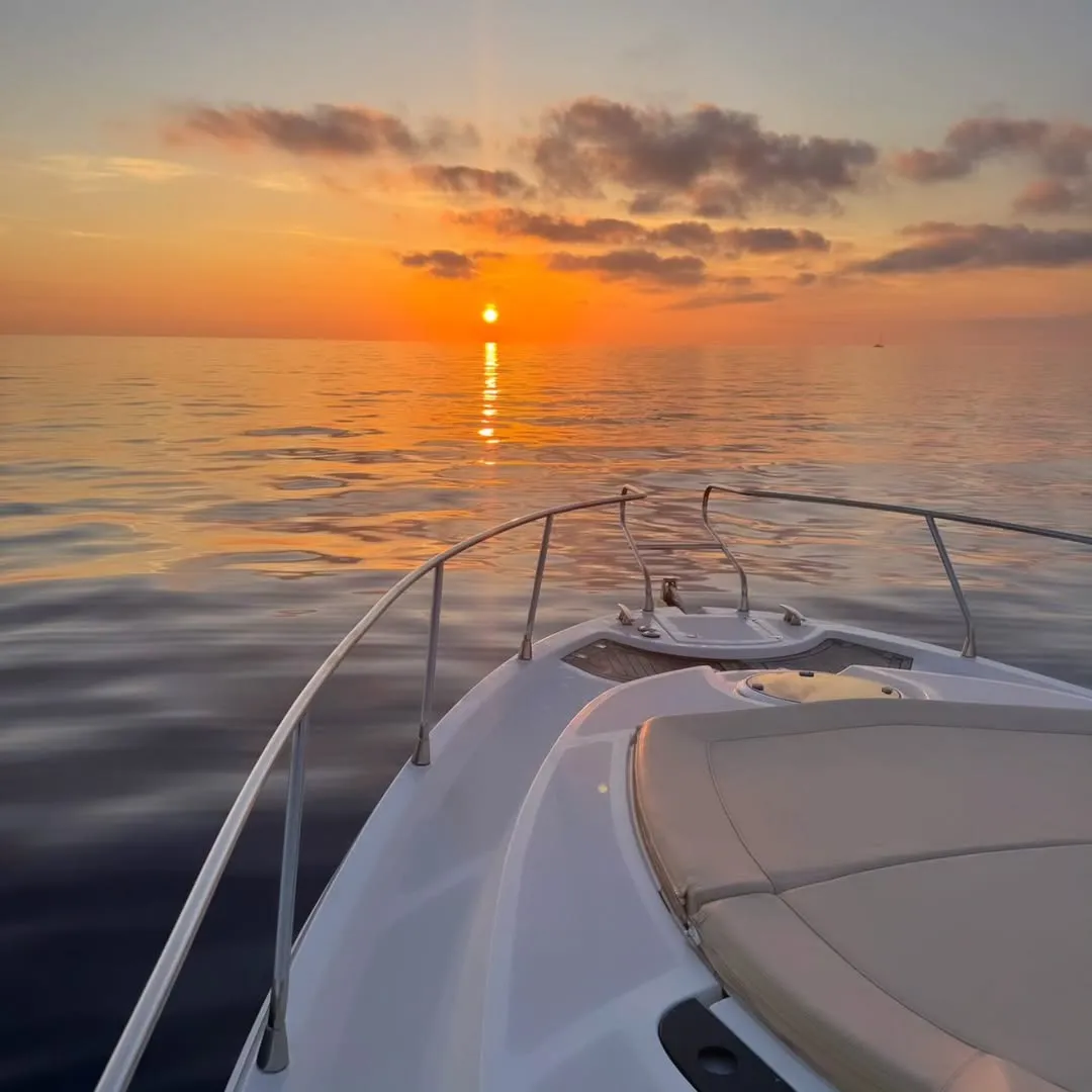The image captures a beautiful sunset scene from the front of a boat on calm waters. The vibrant orange and yellow hues of the setting sun are reflected on the water's surface, creating a visually appealing gradient. The composition places the boat in the foreground, leading the eye towards the expansive horizon and the bright sun.