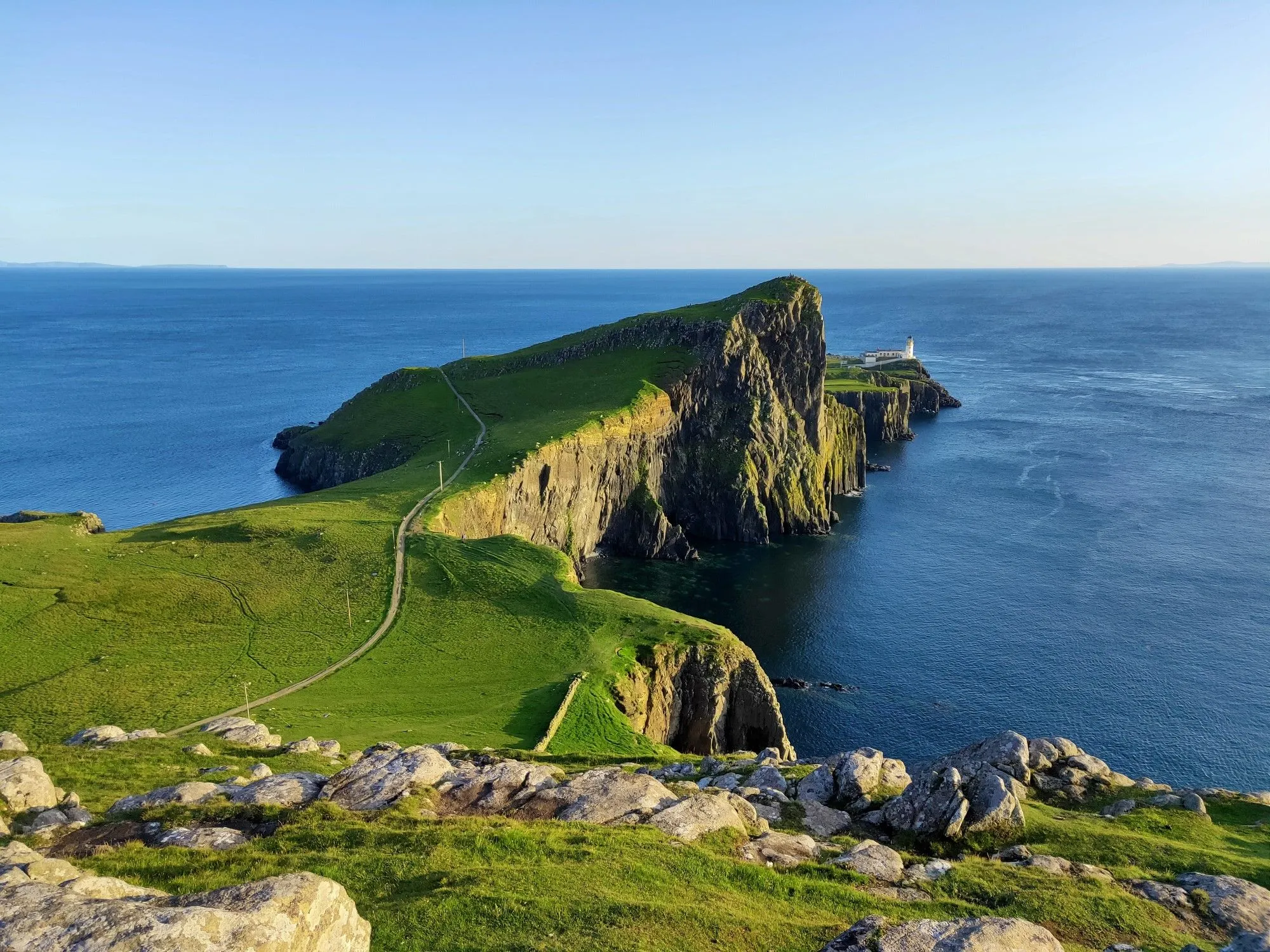 A breathtaking wide shot of a dramatic coastal landscape featuring a verdant peninsula with steep cliffs, leading to a prominent white lighthouse at its end. A winding path traverses the green terrain, bordered by the deep blue sea on both sides under a clear sky. Rocky elements are visible in the immediate foreground, adding depth to the scene.