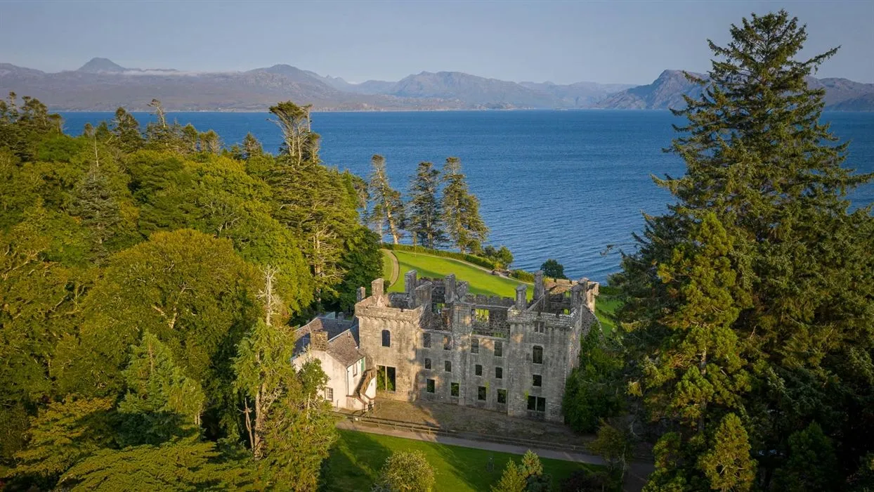 An aerial view of a historic stone castle nestled on a grassy hillside overlooking a large body of water, possibly a loch or sea, with mountains in the background under a clear sky. The castle appears to be in a state of ruin or disuse, adding to its historic charm.