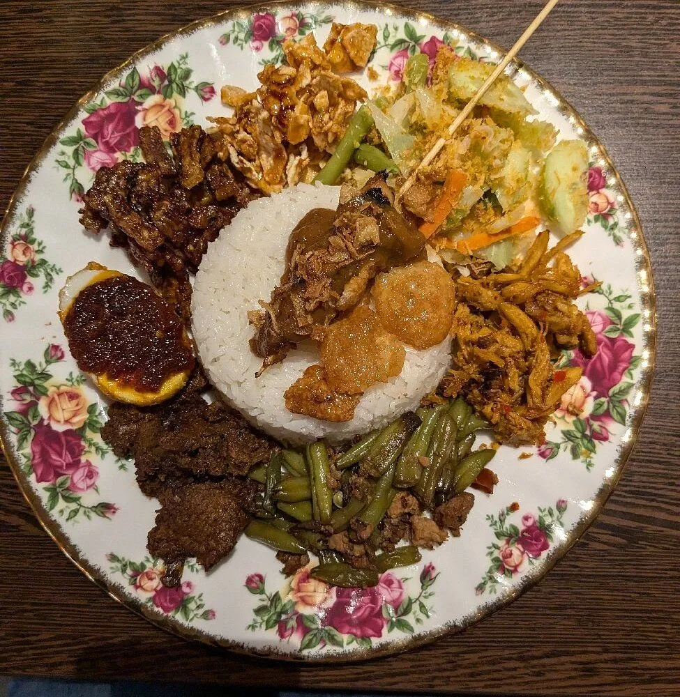 A close-up, top-down view of a traditional Indonesian Nasi Campur dish, featuring a mound of white rice surrounded by various flavorful components including meats, vegetables, a boiled egg with chili sauce, and crispy elements, all presented on a floral-patterned plate.