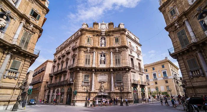The image prominently features a historic Baroque building, likely in a city square, under a clear blue sky. The architectural details are rich and well-defined, capturing the ornate design of the structure. Several people are visible at the base of the building, providing a sense of scale and liveliness to the urban scene.